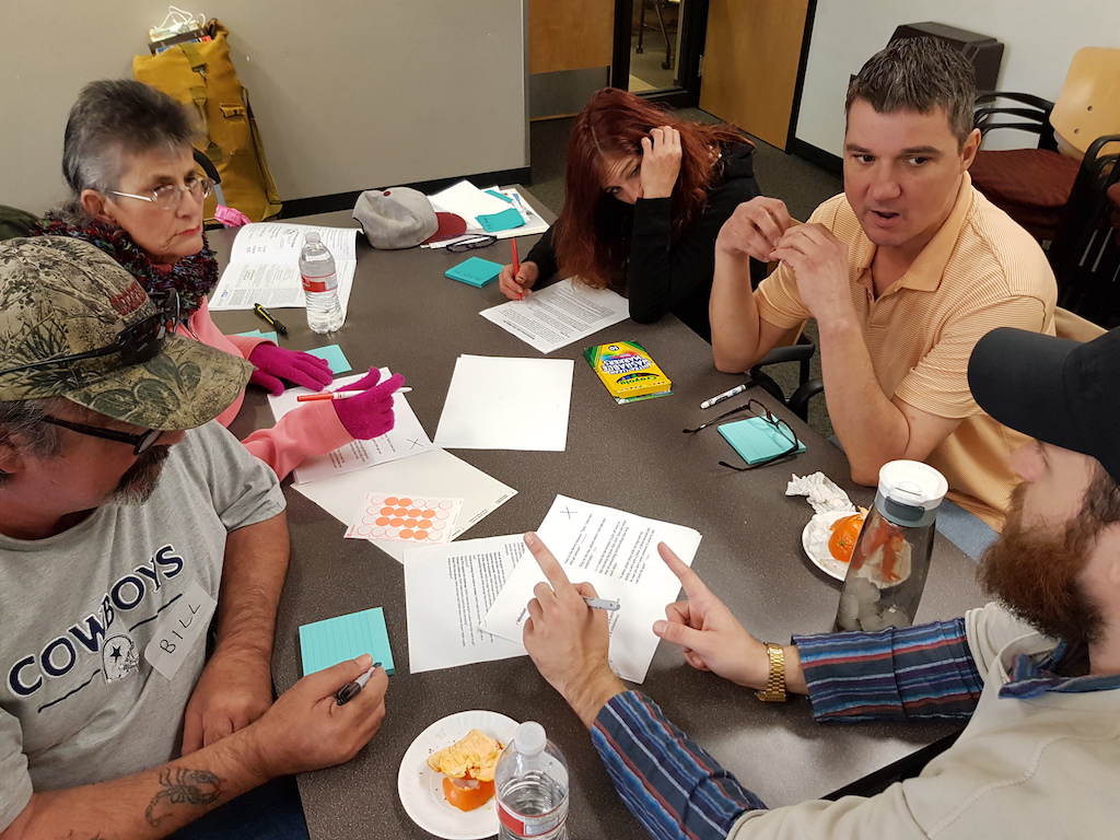 A group of five people sit at a table together, talking. The table is covered in papers, sticky notes, markers, waterbottles, and food.